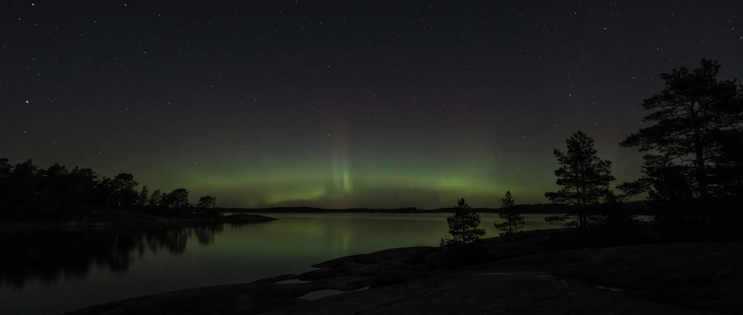 Stilla vik i skärgården en natt med svagt grönt norrsken över horisonten.